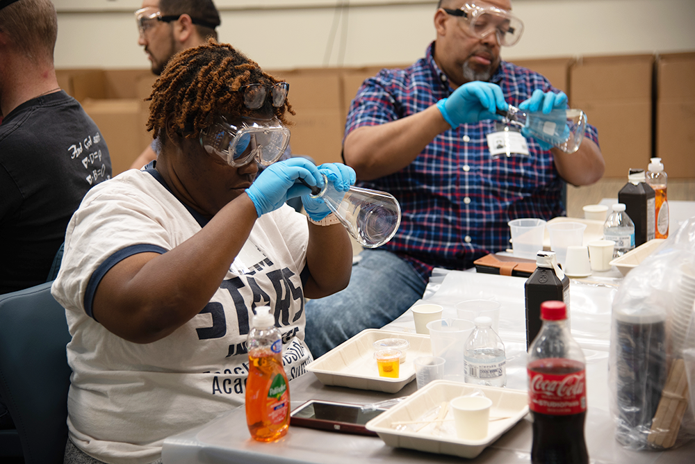 Teachers at a seminar wear safety glasses and look into glass flasks while sitting at a table of experiment supplies. 